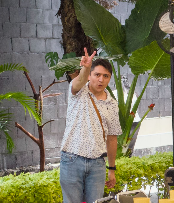 Man in a light patterned shirt and jeans makes a peace sign toward the camera in a garden backdrop.